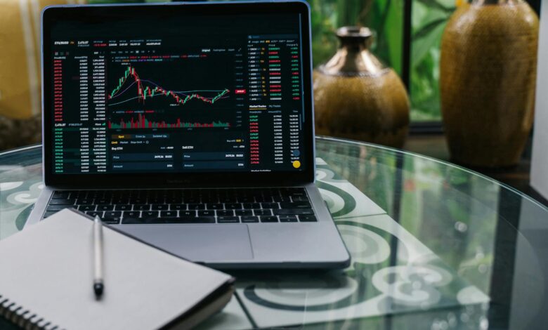 Laptop displaying cryptocurrency stocks and graphs on a glass table with a notepad.